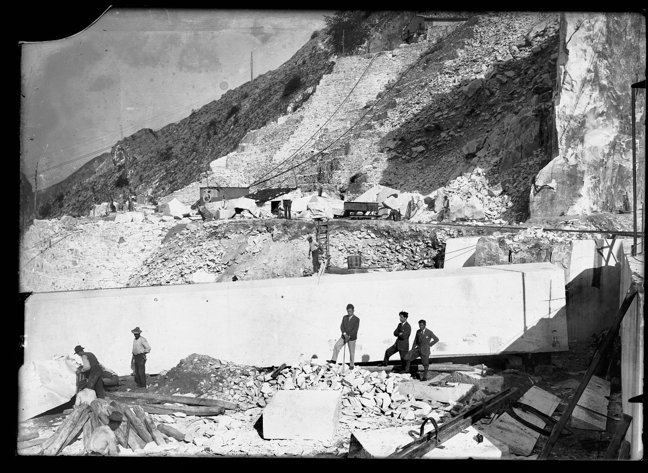 Three men observe the work of several excavators in the Apuan Alps. Behind them is an enormous block of marble already squared off and ready to be transported to the valley by A. Corsini