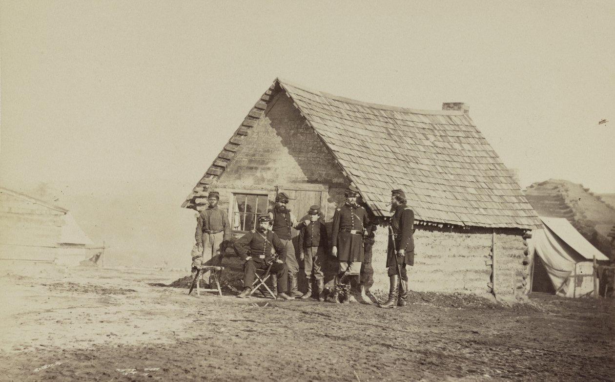 A group of soldiers, and two young men, one an African American, stand outside of log cabin quarters by American Photographer