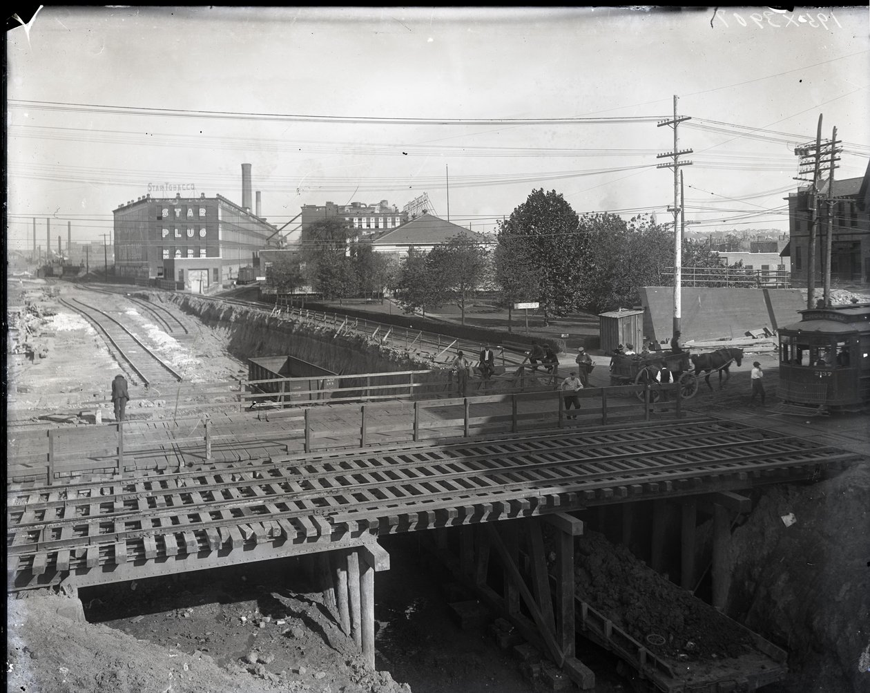 Horizontal, Black and White Photograph Showing a Wooden Bridge and Trolley Tracks over Partially Constructed Railroad Tracks. A Streetcar Marked Chouteau 2137 is Starting to Cross the Bridge on the Far Right. In the Distance Factory Buildings Reading Star by Charles Clement Holt