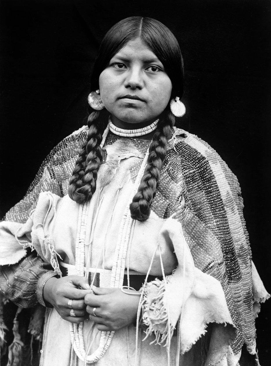 Cayuse woman, half-length portrait, standing, facing front, braids, shell disk earrings, shell bead choker and shell beads around neck, beaded buckskin dress with beaded belt c. 1910, photo Edward S. Curtis by Edward Sheriff Curtis