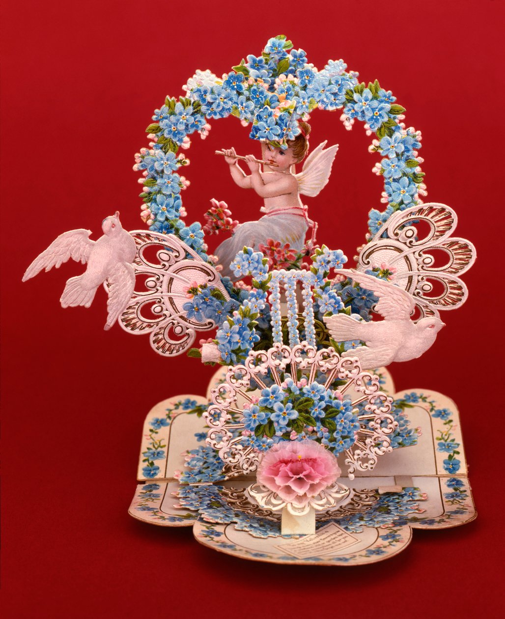 A pop-up card a winged cherub playing a flute under a heart shaped arch of forget-me-nots by English School