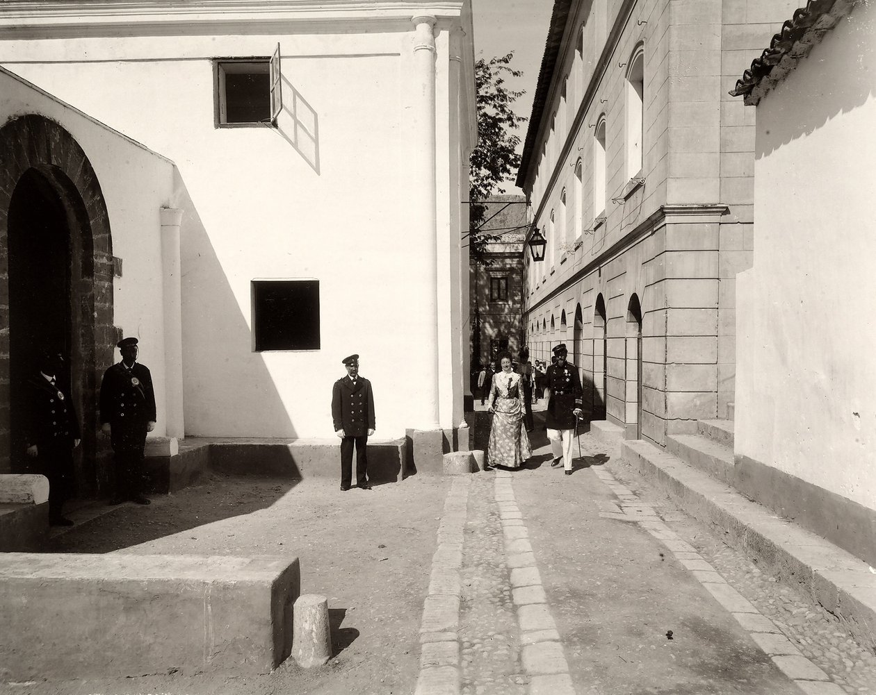 Men in uniform accompanying an elegantly dressed woman towards the church of the farm of Lo Zucco, Sicily by Eugenio Interguglielmi