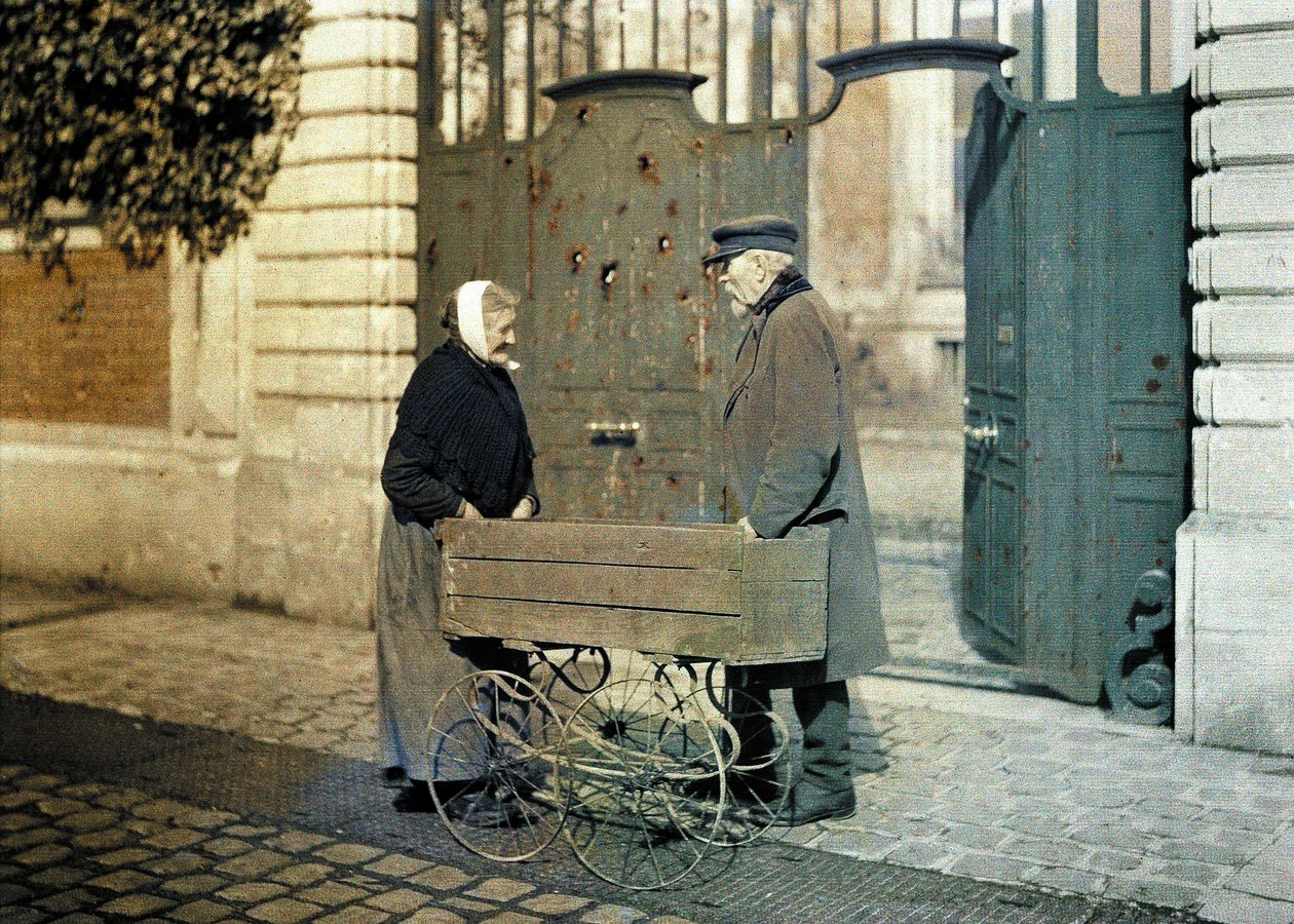 Two senior citizens of Reims, an old woman and Mr. Reiser, standing in front of a gate with many bullet holes, Reims, Marne, France, 1917 (autochrome) by Fernand Cuville