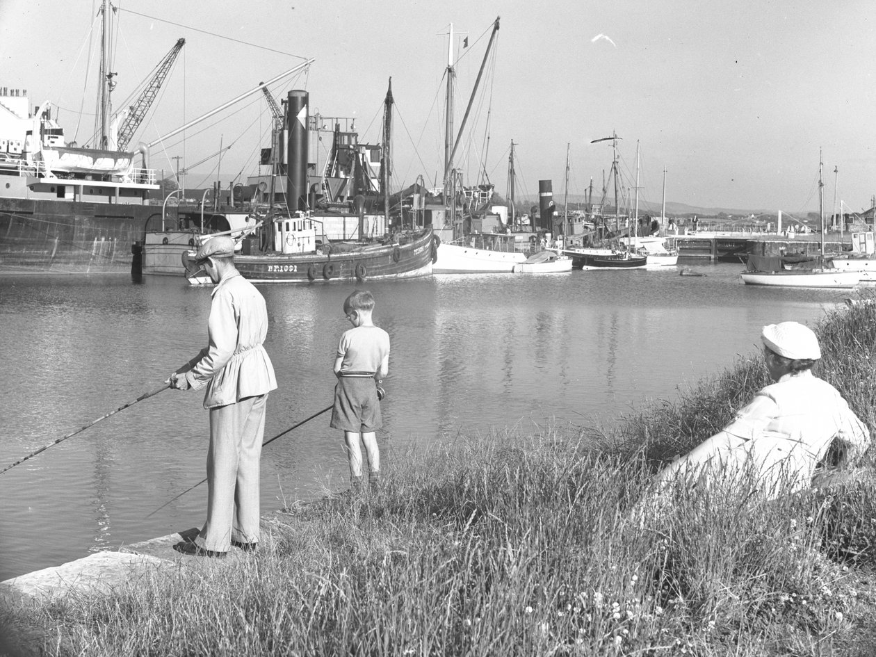 A man and boy fishing in Glasson Dock, lady looking on, various boats moored by Joseph Hardman