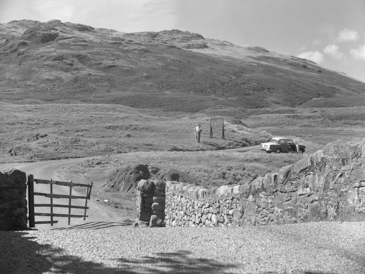 View of the road passing over Hard Knott Pass, two people standing near a road sign and a parked car on grass verge by Joseph Hardman