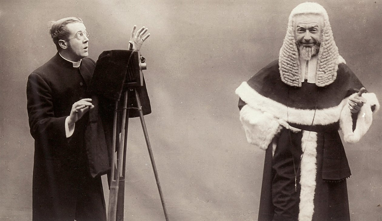 A man in a cassock aims the lens of his camera at a man dressed like a pope by Rotary Photographic Co Ltd