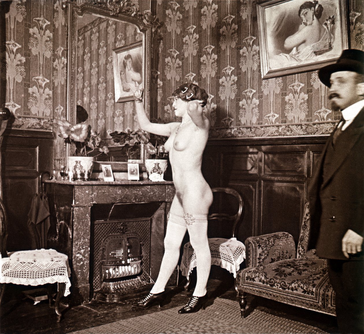Enigmatic photograph showing a young woman completely naked (except for her shoes and stockings) recoiffing her hair in front of a mirror in the presence of a man dressed and heated with a hat. One might think it was a photo taken in a brothel, but this hy by Unbekannter Künstler