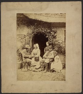 Group seated outside open doorway: two men in hats, woman in bonnet, two young girls - one standing