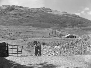 View of the road passing over Hard Knott Pass, two people standing near a road sign and a parked car on grass verge by Joseph Hardman
