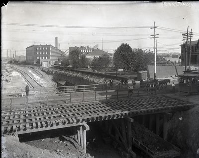 Horizontal, Black and White Photograph Showing a Wooden Bridge and Trolley Tracks over Partially Constructed Railroad Tracks. A Streetcar Marked Chouteau 2137 is Starting to Cross the Bridge on the Far Right. In the Distance Factory Buildings Reading Star by Charles Clement Holt