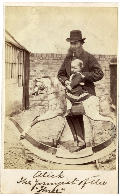 Young girl on a rocking horse in the garden, her father beside by English Photographer