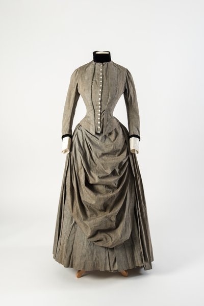 Grey cotton dress with draped apron front and black velvet trimming at collar and cuffs, late 1880s by English School