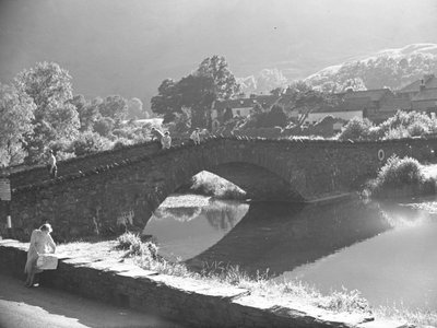 Woman sitting on wall studying map with river and stone bridge in background by Joseph Hardman