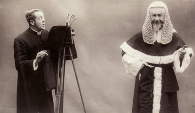 A man in a cassock aims the lens of his camera at a man dressed like a pope by Rotary Photographic Co Ltd