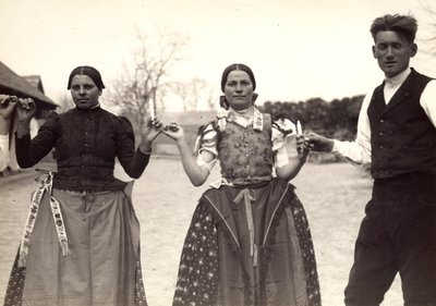 Two ladies and a man, in Hungarian traditional dresses, are holding hands in a dance, holding some candles in their hands by Rudolf Balogh