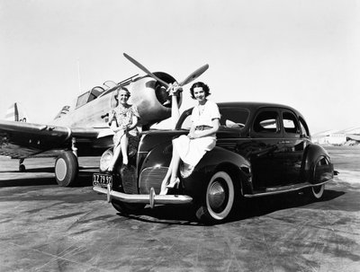 California: 1939 Two women sit on the fenders of a 1939 Lincoln Zephyr sedan at an airport with a single engine plane parked behind them by Unbekannt