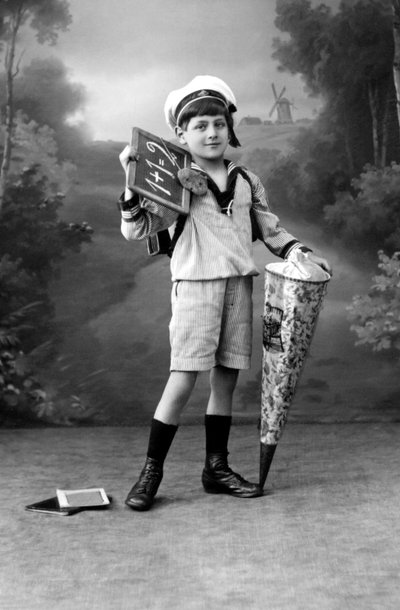 First day of school. A young boy in uniform posing with a slate showing the addition 1+1 = 2 and a surprise pouch. Photography around 1910. by Ferdinand Georg Waldmuller