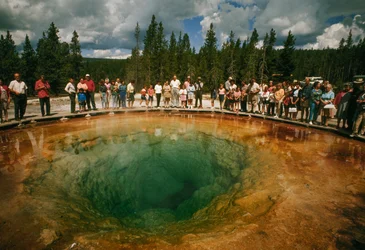 Park visitors gather around a blue-green, blossom-shaped thermal pool. Yellowstone National Park