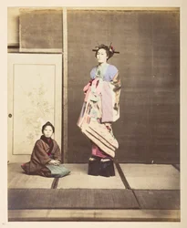 Full-length standing studio portrait of a young woman in elevated shoes, her young attendant kneeling on the floor beside her