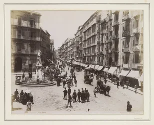 View of Via Roma in Naples with a statue, shops, horse-drawn carriages and passers-by