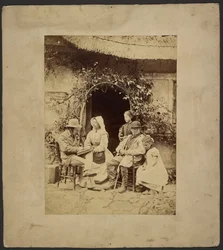 Group seated outside open doorway: two men in hats, woman in bonnet, two young girls - one standing