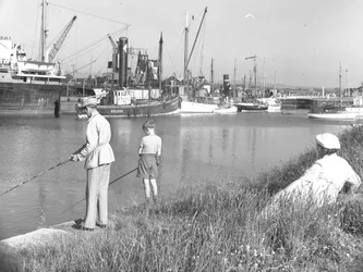 A man and boy fishing in Glasson Dock as a lady seated behind looks on, various boats moored up