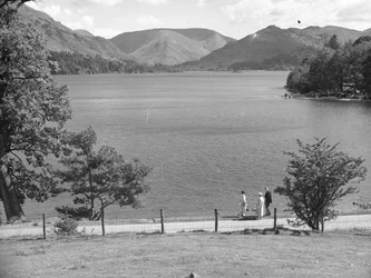 A man, lady and possibly their daughter stroll along the edge of a lake, hills in the background