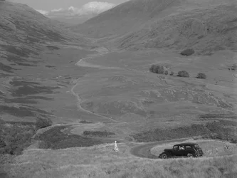 A woman stands overlooking an open valley, with a parked car nearby