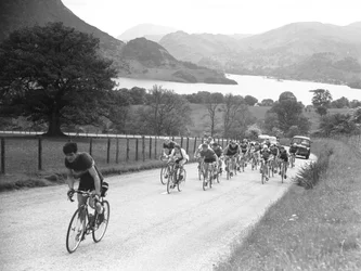 Group of cyclists heading up a road followed by cars, lake and hills in background