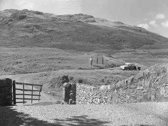 View of the road passing over Hard Knott Pass, two people standing near a road sign and a parked car on grass verge
