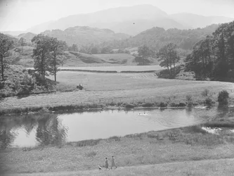 Women pushing baby in pushchair on nearside of river through a meadow with people swimming in river and farmer working tractor in fields beyond and fells in background
