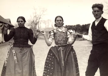 Two ladies and a man, in Hungarian traditional dresses, are holding hands in a dance, holding some candles in their hands
