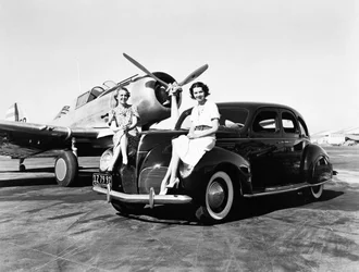 California: 1939 Two women sit on the fenders of a 1939 Lincoln Zephyr sedan at an airport with a single engine plane parked behind them
