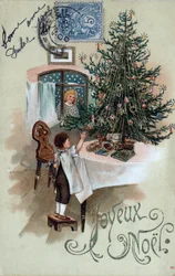 Christmas greeting card: a child in front of a Christmas tree poses on a table, observed by an angel in the window