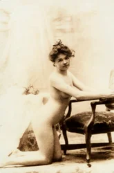 Nude portrait of a young woman kneeling in front of a small armchair