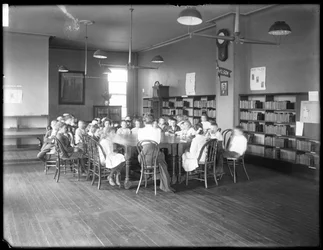 Children listen to a woman in the reading room of an unidentified branch of the Queens Borough Public Library, c. 1910