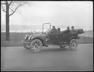 Three unidentified people and two Chow dogs driving in a convertible car along a river or lake