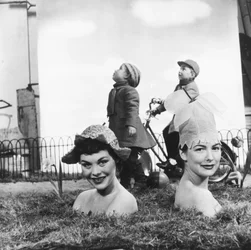 Women wearing flower-shaped hats, with bodies hidden in the ground giving the illusion of emerging from the earth